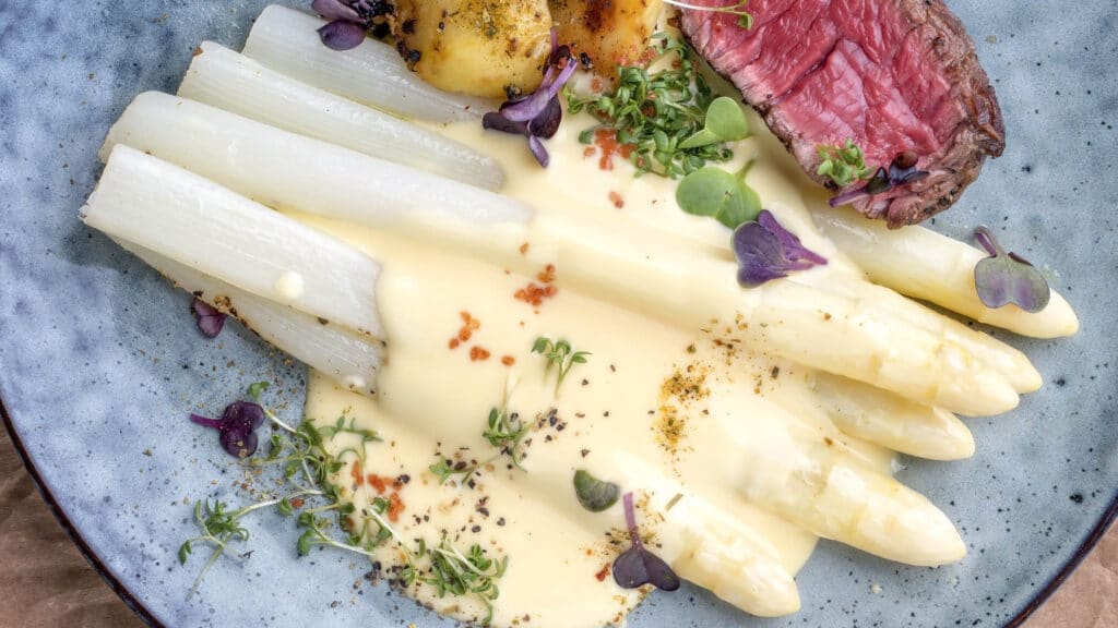 Weiße Spargel mit Sauce Hollandaise und Fleisch und Kartoffeln auf einem blauen Teller